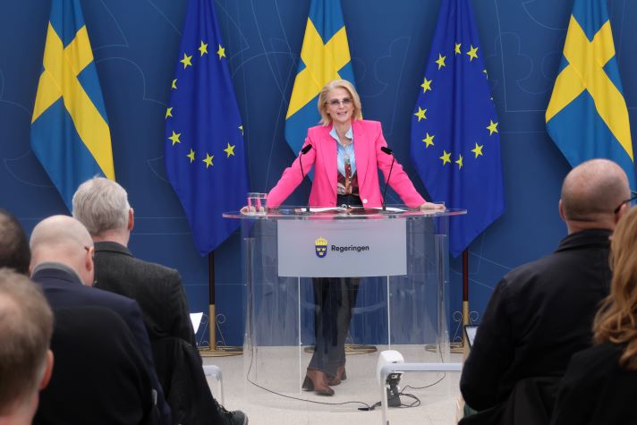 Minister for Finance, Elisabeth Svantesson, is standing on a stage against a backdrop of Swedish and EU flags. 