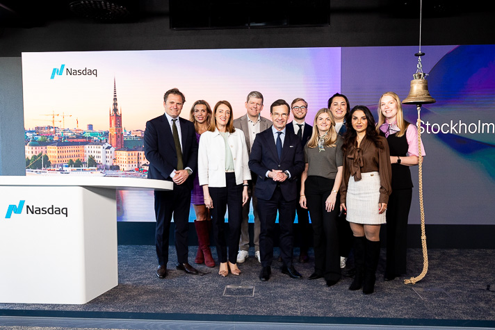 Ulf Kristersson and Roberta Metsola are standing with eight others in a group photo. To the left of the group, the Nasdaq stock exchange logo is visible, and to the right, the exchange’s bell hangs from a rope.