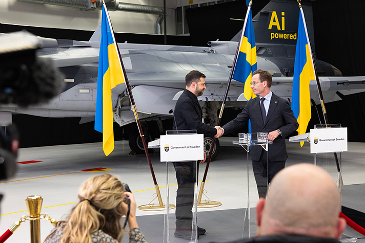 Volodymyr Zelensky and Ulf Kristersson stand at a press conference and shake hands. In the background JAS Gripen and Ukrainian and Swedish flags. Journalists are visible in the foreground.