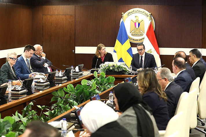 A meeting room where delegates are seated around a large conference table. Swedish and Egyptian flags stand behind two speakers at the front, and plants decorate the centre of the table