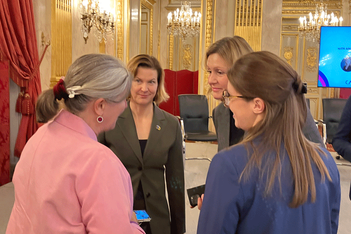 The Swedish Minister for Gender Equality and the President of the French National Assembly are talking with the French Minister for Equality and another participant. In the background there are golden details on the wall and red curtains.