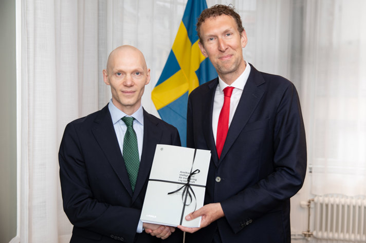 Two men looking into the camera, shaking hands. They have a printed copy of an application in their hands. Swedish flag in background, formal wear.